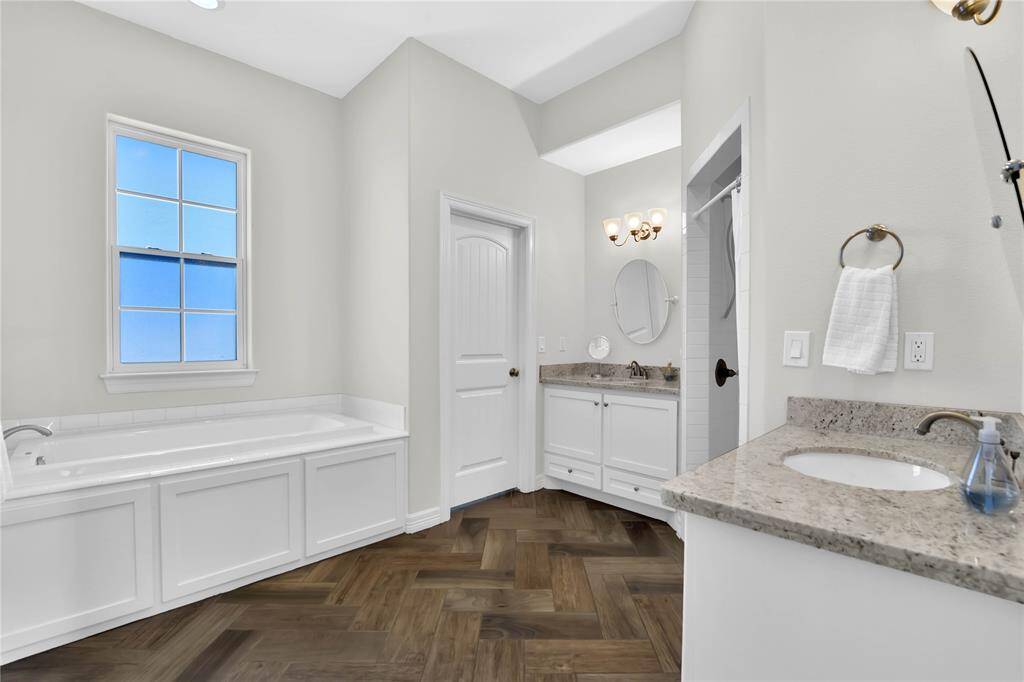 This spacious bathroom features a large soaking tub, dual sinks with granite countertops, a separate shower area, and elegant wood-patterned flooring, all complemented by soft, neutral walls and ample natural light.