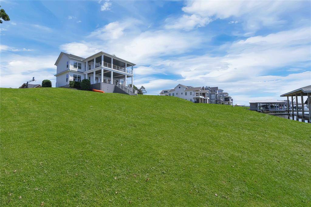 This photo features a spacious, modern two-story home with a large covered patio, situated on a green hillside. The property offers scenic water views and is part of a neighborhood with similar waterfront homes.