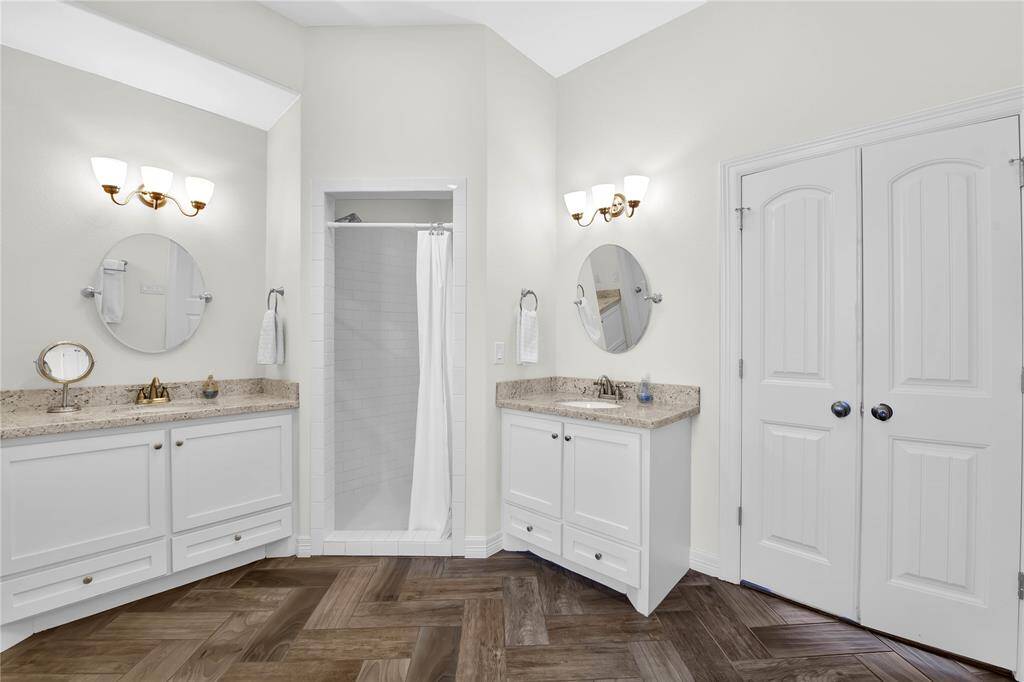 This bathroom features dual vanities with granite countertops, elegant lighting, a central walk-in shower, and a herringbone patterned wooden floor.