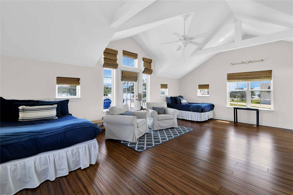 This spacious bedroom features high vaulted ceilings, abundant natural light from multiple windows, and elegant wood flooring. It includes two beds, cozy seating with armchairs, and a stylish area rug, creating a comfortable and inviting atmosphere.