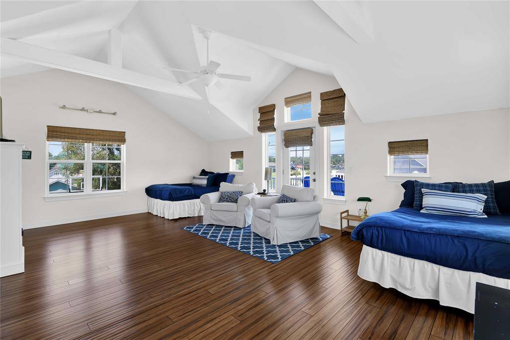 This spacious, bright room features vaulted ceilings with white beams, hardwood flooring, and large windows with wooden blinds. It includes two cozy beds, a comfortable seating area, and a neutral color palette, creating a welcoming atmosphere.