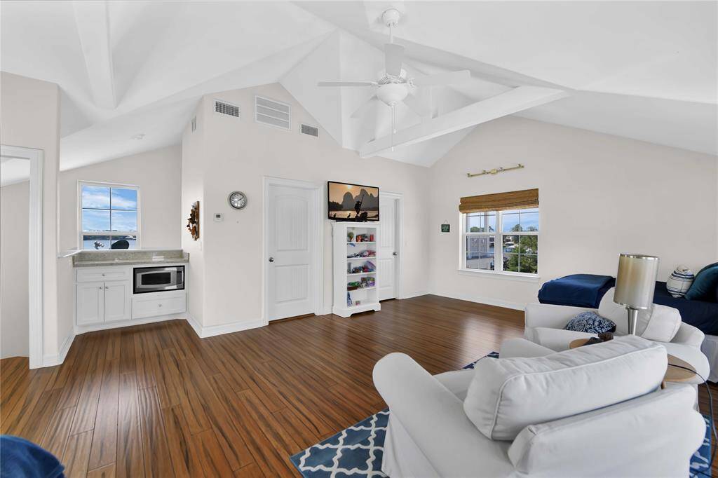 Bright, open living space with vaulted ceilings and wood flooring. Features include large windows, a cozy sitting area, and a small kitchenette with a microwave. Perfect for relaxation with ample natural light.