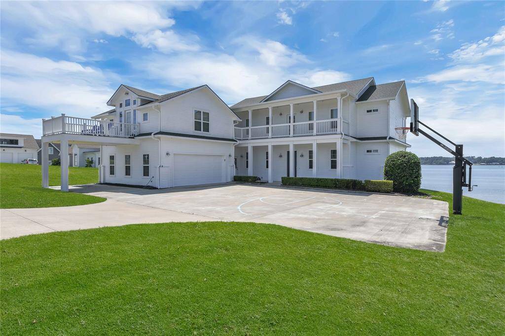 Spacious two-story waterfront home featuring a large driveway with basketball hoop, wraparound balcony, and lush green lawn. Ideal for enjoying scenic views and outdoor activities.