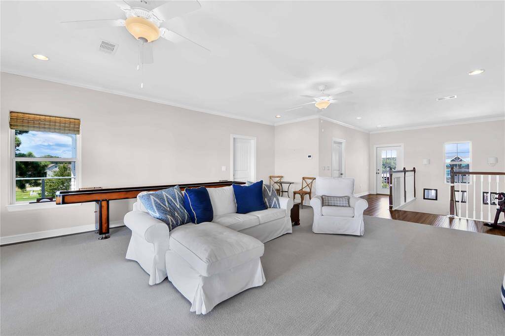 This spacious, bright living area features neutral-toned walls and carpet, with a cozy sofa and armchair accented by blue pillows. Natural light floods the room through large windows, and there's a shuffleboard table for entertainment. The space has recessed lighting and ceiling fans, creating a comfortable atmosphere.