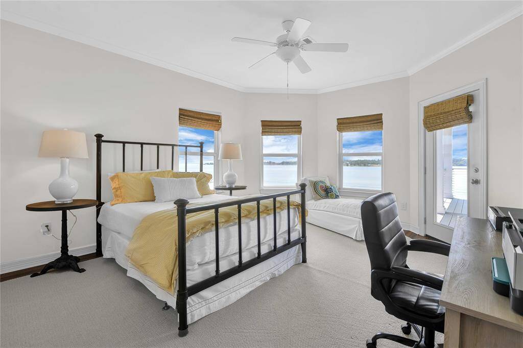 Bright bedroom with a cozy bed, desk, and stunning lake views through multiple windows. Features a ceiling fan and neutral decor for a serene atmosphere.