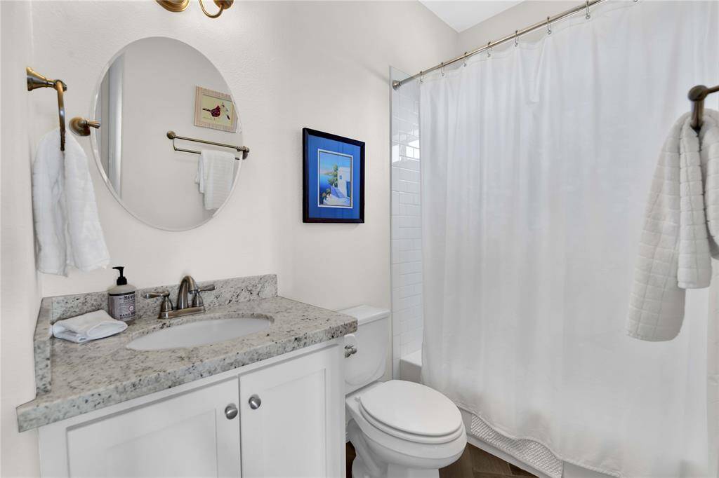 This bathroom features a modern design with a granite countertop, under-mount sink, and white cabinetry. It includes a shower-tub combo with a white curtain, a round mirror above the sink, and tasteful wall art. The space is bright and inviting, ideal for a comfortable home setting.