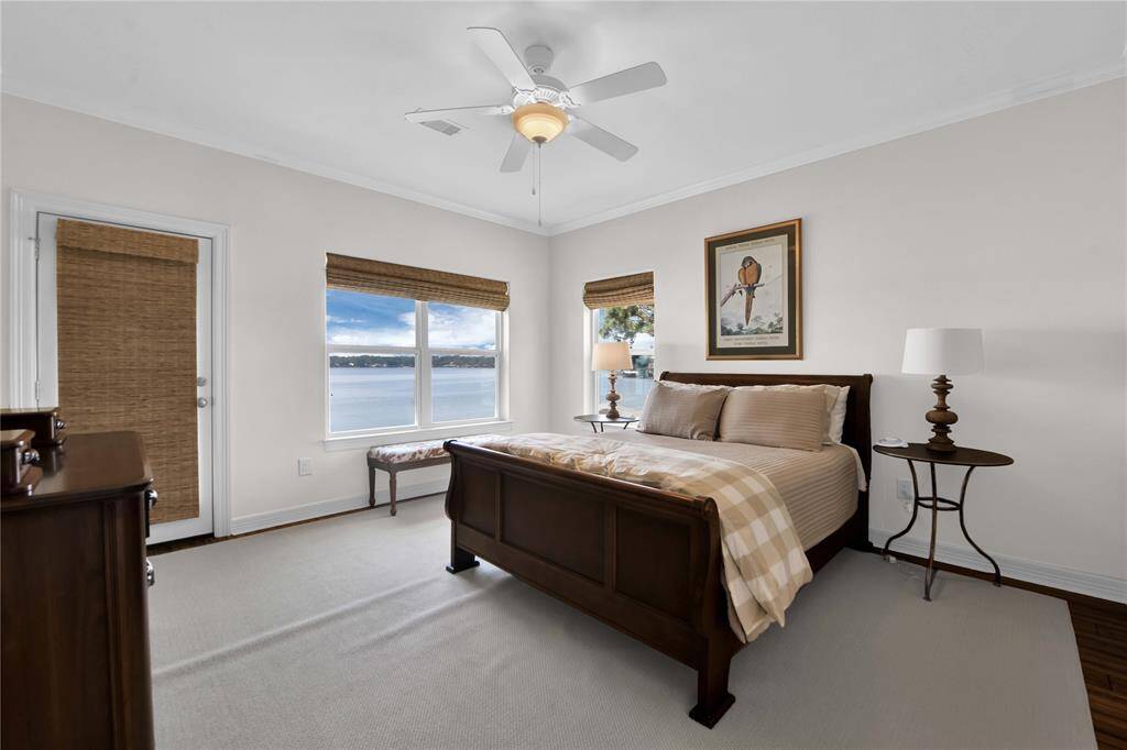 This bedroom features a cozy, elegant setup with natural light filling the room through large windows offering a stunning water view. Neutral tones and a ceiling fan add to the serene atmosphere.