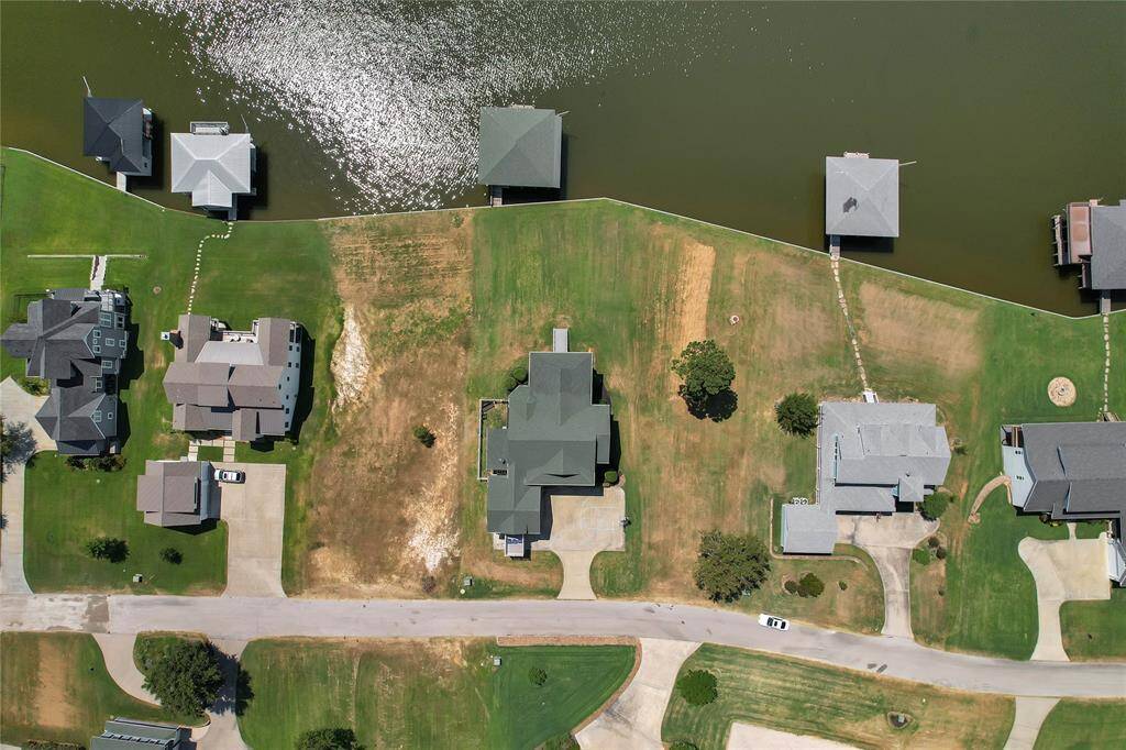 This aerial photo shows a row of waterfront homes with private docks along a calm body of water. The properties have spacious yards, and there's a street with driveways providing access to each home. Ideal for buyers seeking peaceful waterfront living.
