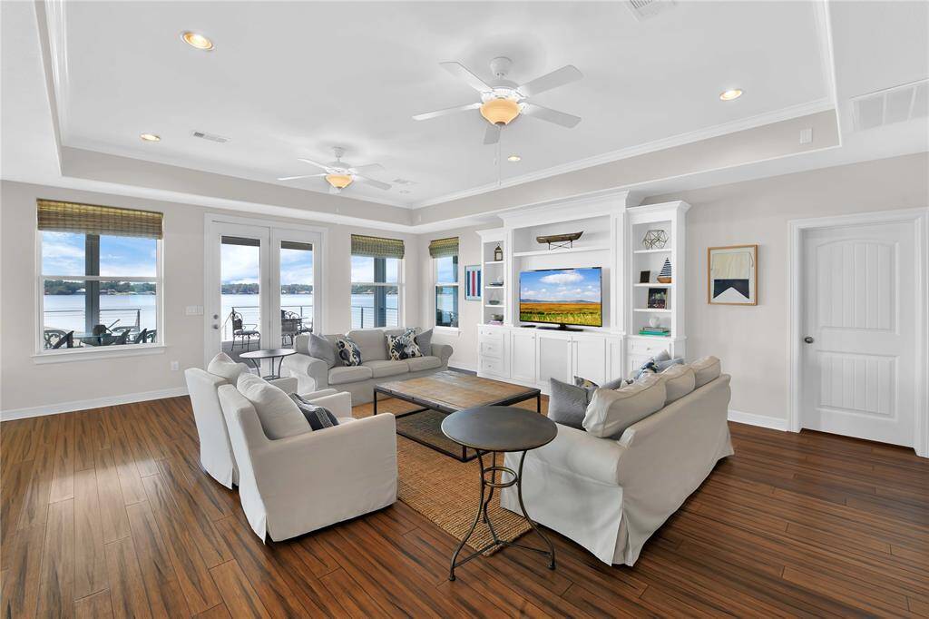 Bright and spacious living room with hardwood floors and tray ceiling. Features large windows and glass doors offering stunning water views. Includes built-in shelving and a cozy seating area.