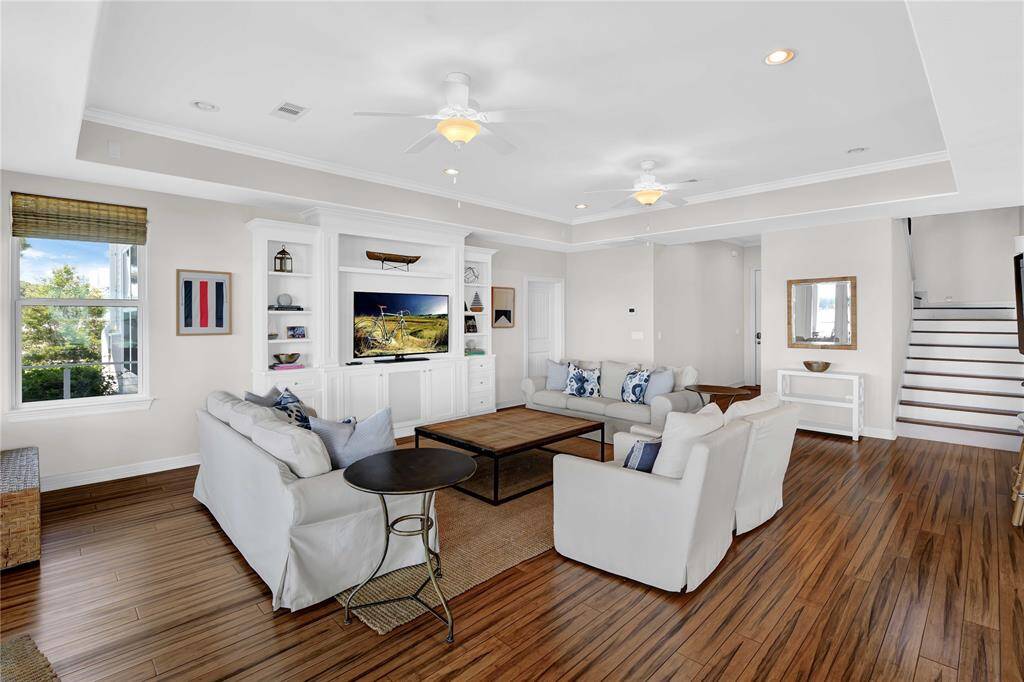 This spacious and inviting living room features elegant wood flooring, a cozy seating area with plush sofas, and a built-in entertainment center. Ample natural light streams through a large window, and the tasteful decor enhances the room's warm and welcoming ambiance.