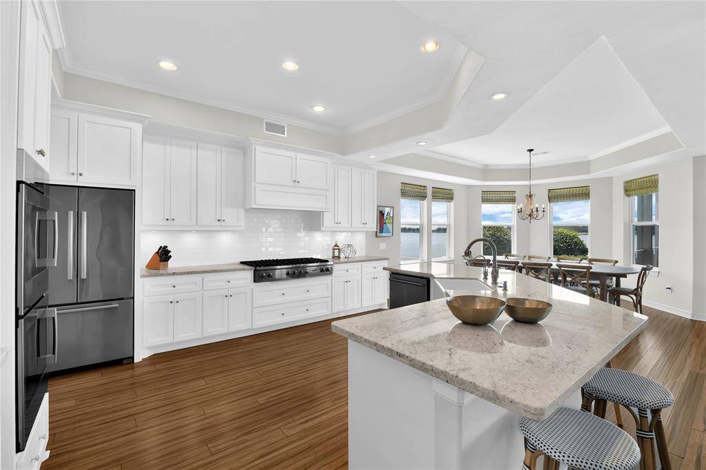 This photo shows a bright, spacious kitchen with white cabinetry, stainless steel appliances, and a large island with seating. The kitchen opens to a dining area with large windows offering scenic views, filling the space with natural light.