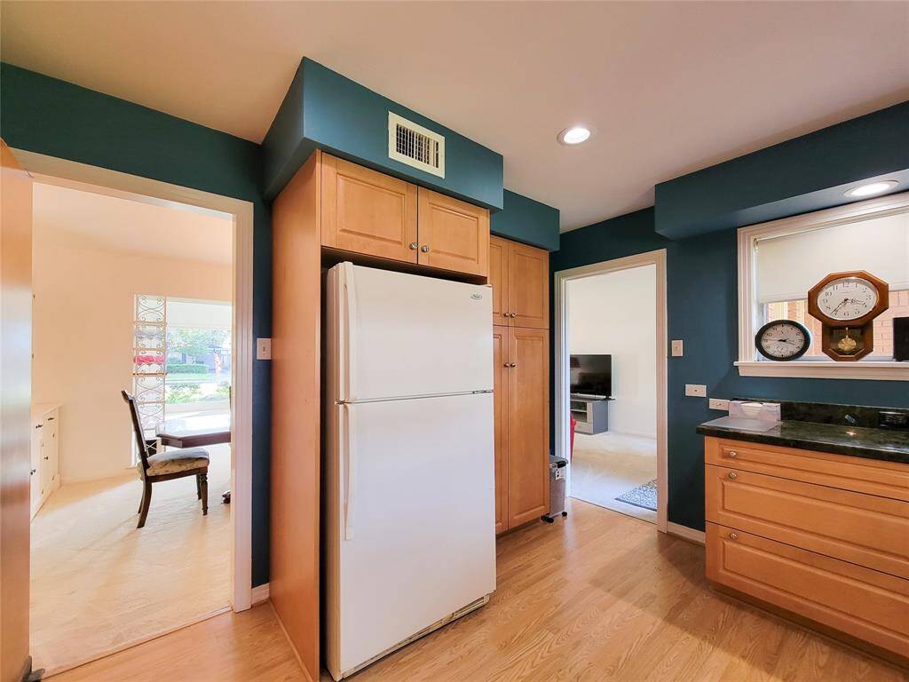 This cozy living room features neutral carpeting, a mix of classic and floral-patterned furniture, and a large TV. Natural light filters through sheer curtains on the double doors, enhancing the room's warm ambiance. An adjoining area is visible, giving a sense of spaciousness and connectivity.