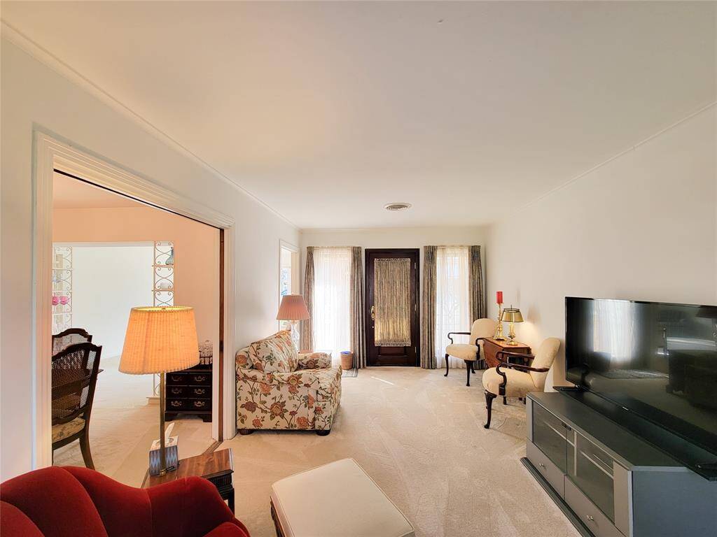 This cozy living room features neutral walls, plush carpeting, and ample natural light from large windows. It’s furnished with a floral-patterned armchair, a red accent chair, and classic wooden side tables, creating a warm and inviting atmosphere.