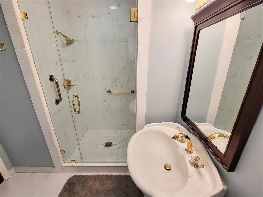 This bathroom features a modern, glass-enclosed shower with elegant gold fixtures and a classic white pedestal sink, complemented by a stylish mirror.