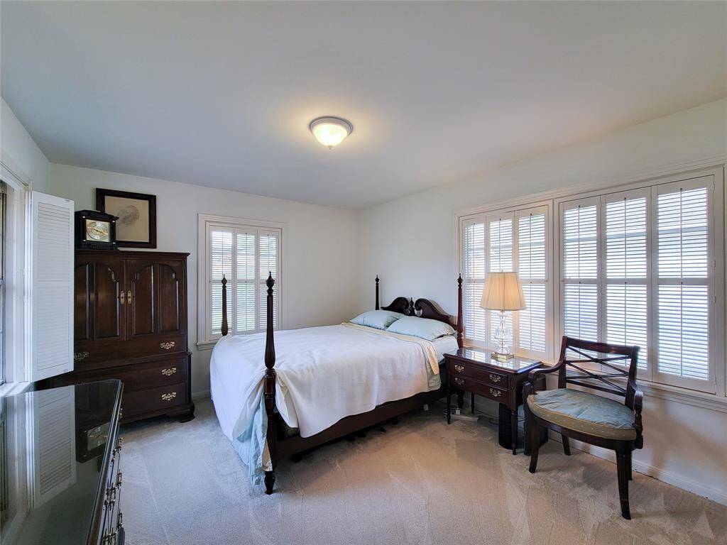 Bright and inviting bedroom with classic furniture, featuring a comfortable bed, large dresser with mirror, and a mounted TV. Ample natural light from two windows enhances the cozy atmosphere.