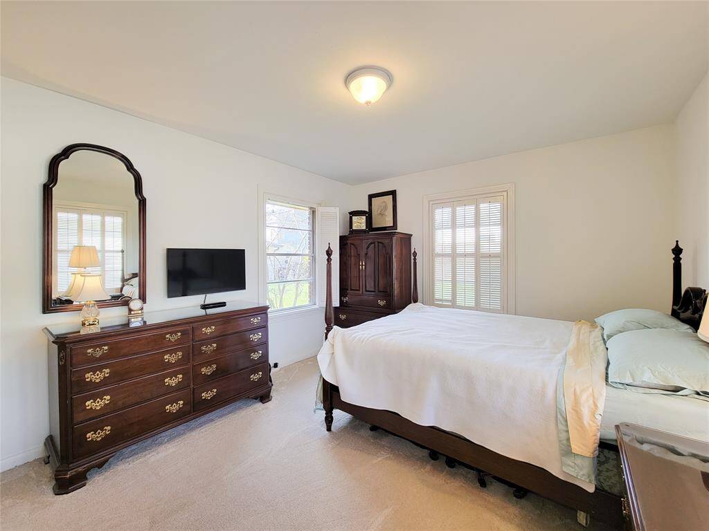 This photo showcases a cozy bedroom with  corner windows with shutters, and warm lighting. The neutral carpet and walls create a calm atmosphere, and the hallway leads to additional rooms.