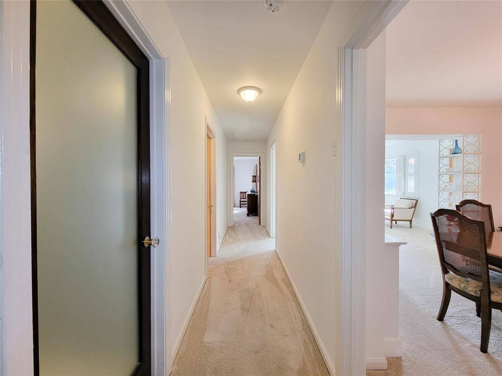 This photo shows a hallway with two frosted glass doors featuring dark wood frames. To the left, there's a doorway leading to a bright bedroom with a bed, wooden furniture, and a window with white shutters. The area is carpeted, providing a cozy feel.