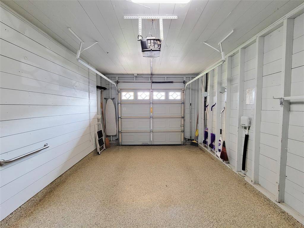 This photo shows a clean, well-organized garage with epoxy flooring and white paneled walls. The space is equipped with a standard garage door, storage hooks, and some cleaning tools neatly arranged, offering a functional and tidy environment.