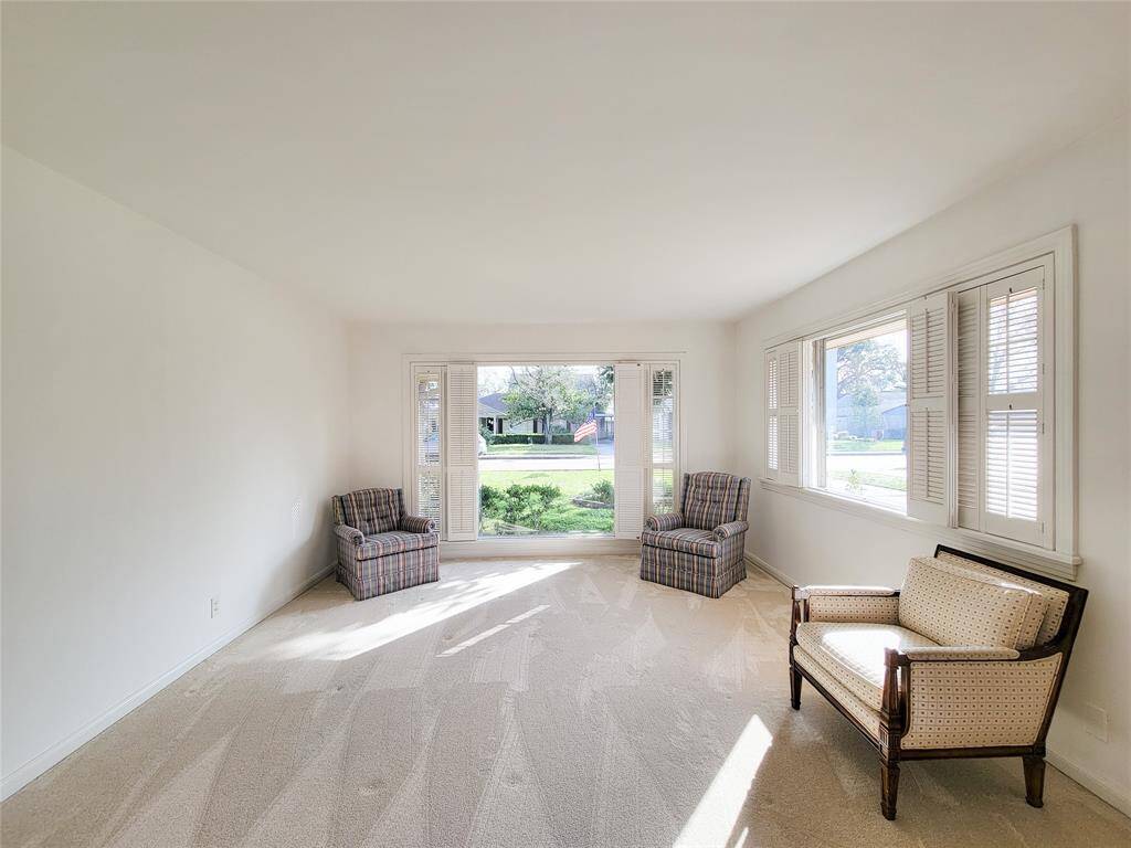 This bright and spacious living room features large picture windows with shutters, allowing plenty of natural light. The neutral-colored carpet and walls create a cozy atmosphere.