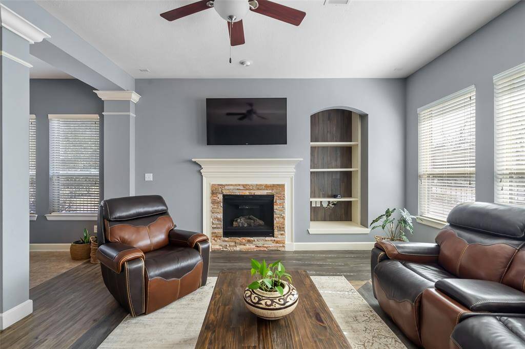 This cozy living room features a modern gas fireplace with a stone surround, built-in shelving, and a wall-mounted TV. It has ample natural light from large windows, complemented by elegant leather seating and a ceiling fan.
