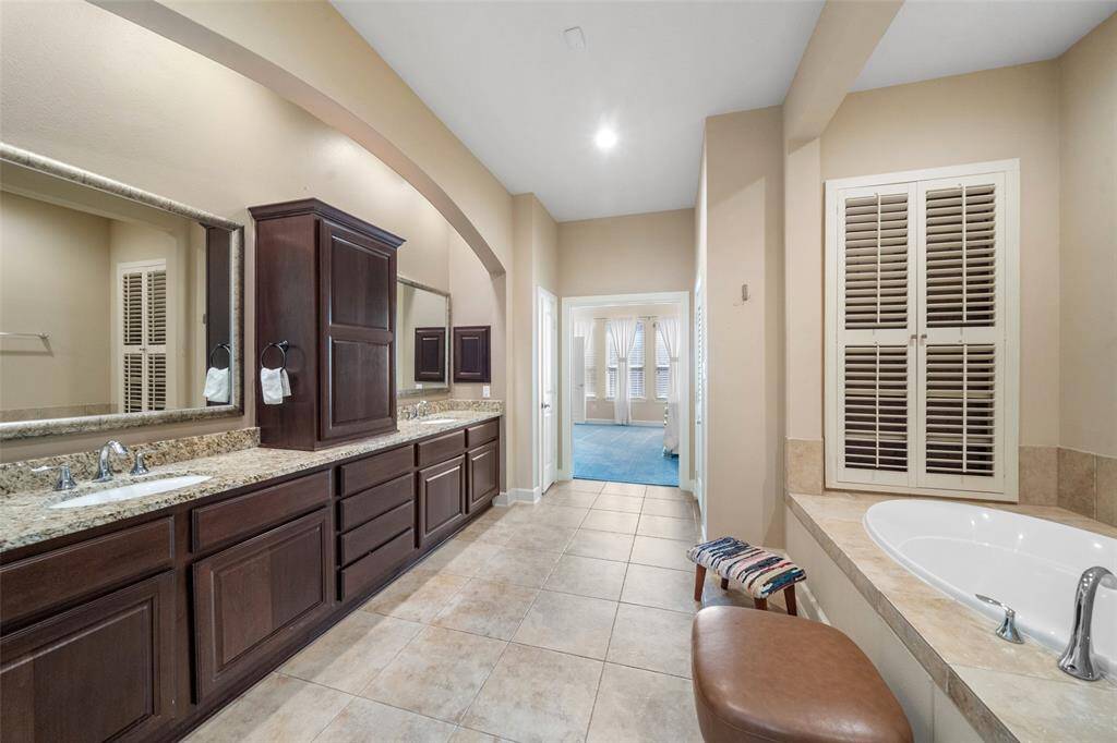 This spacious bathroom features dual granite vanities, ample cabinetry, a built-in bathtub, and elegant tile flooring. An arched entryway leads to a bright room with large windows, enhancing the open feel.