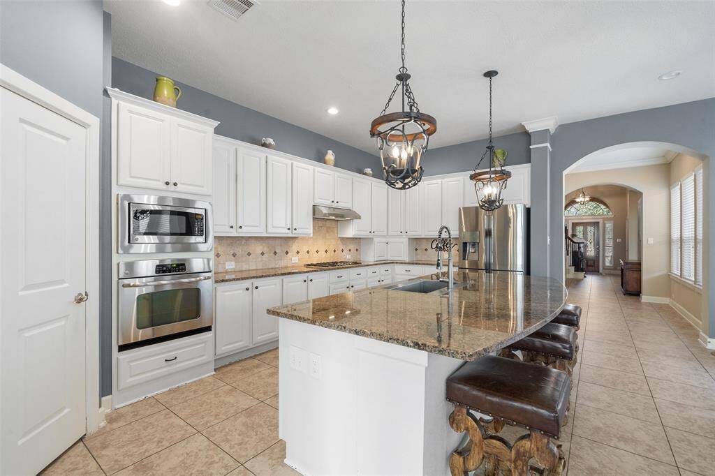 This spacious kitchen features elegant granite countertops, stainless steel appliances, and ample white cabinetry. It has a large island with seating, pendant lighting, and an open layout leading to a bright hallway.