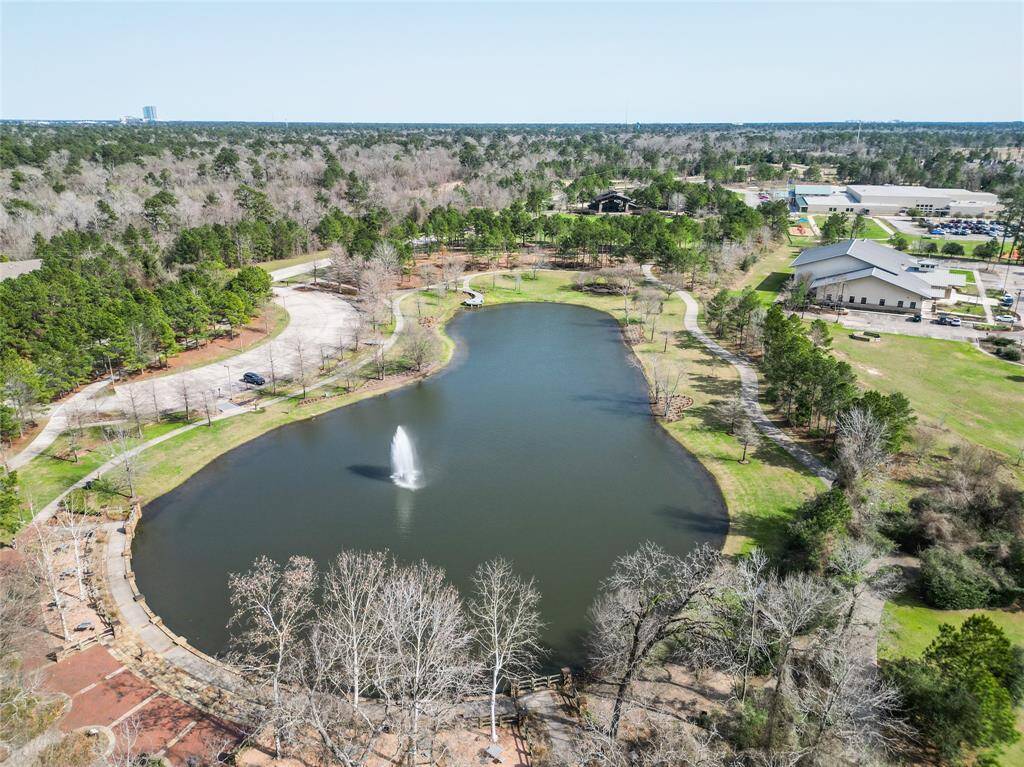 A scenic pond featuring a fountain, surrounded by green spaces and walking paths. Nearby amenities include parking and community buildings, all set against a backdrop of natural woodland.