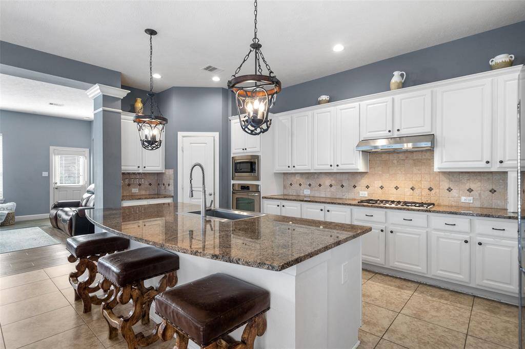 This kitchen features a spacious layout with a large granite island and stylish pendant lighting. It includes ample white cabinetry, stainless steel appliances, and a gas cooktop. The open design connects to an adjacent living area, creating a welcoming space for cooking and entertaining.