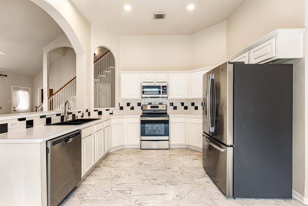 This bright and elegant kitchen features crisp white cabinetry, modern black and white tile backsplash, and sleek stainless steel appliances. The open-concept design is complemented by high ceilings, recessed lighting, and arched openings that create a spacious and airy feel. A stylish black sink and faucet contrast beautifully with the light countertops, while the large layout provides ample storage and prep space. A perfect blend of classic charm and modern convenience.