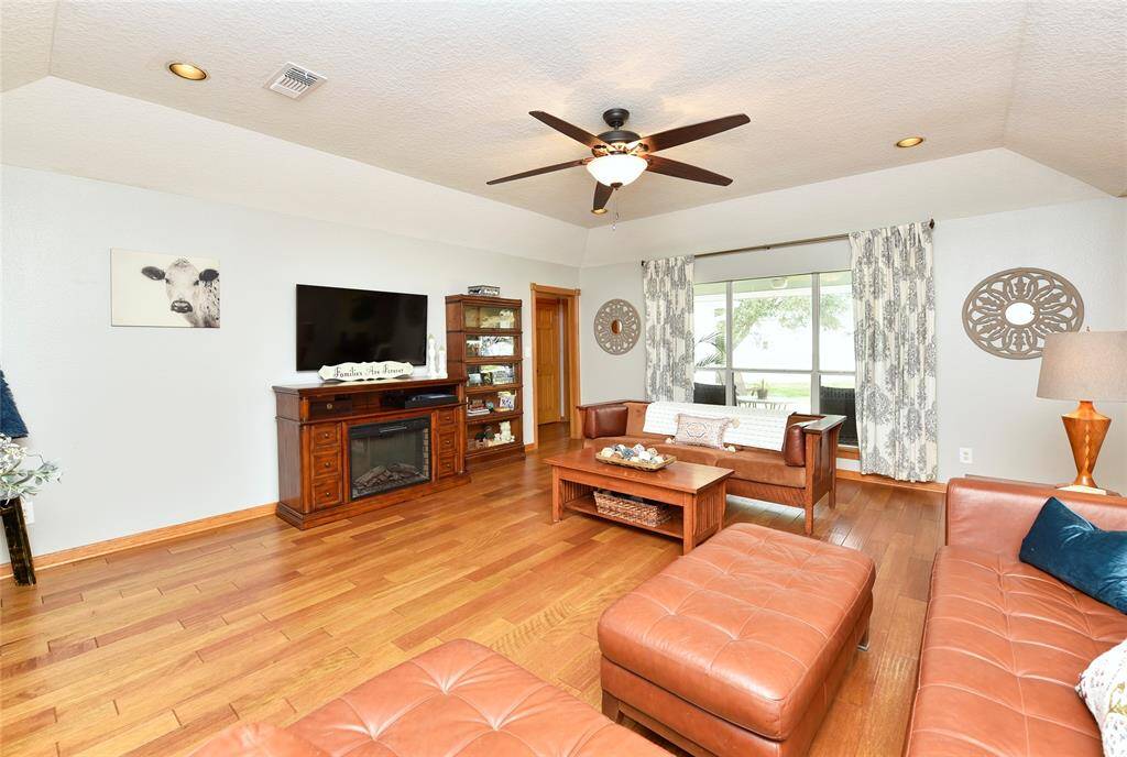 This inviting living room features hardwood floors, cozy leather seating, and a ceiling fan. A large window provides natural light and a view of the outdoors. The space includes a TV above a classic wooden fireplace and tasteful decor, creating a warm, welcoming atmosphere.