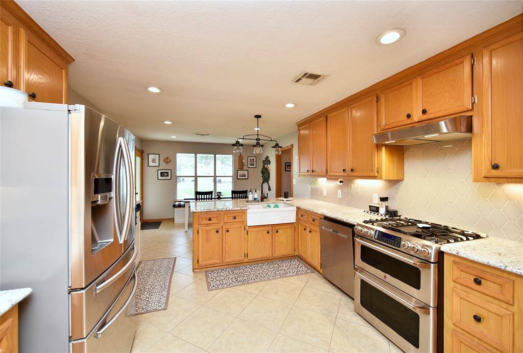This kitchen features warm wood cabinetry, stainless steel appliances, and granite countertops. The space includes a farmhouse sink, a gas range, and ample storage. A dining area with large windows provides natural light, creating an inviting atmosphere.
