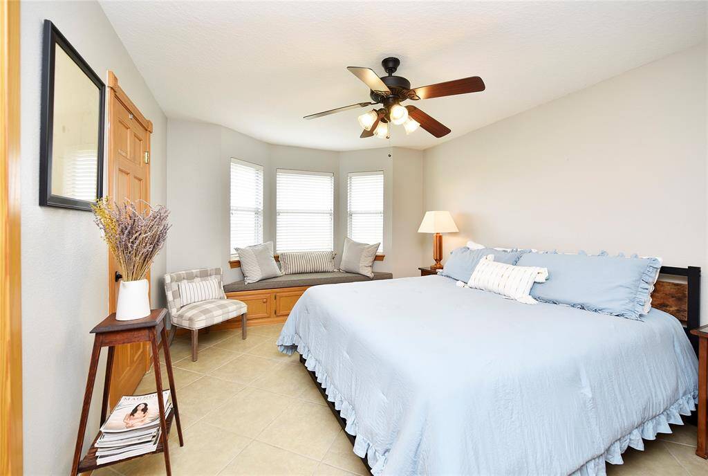 This bedroom features a cozy atmosphere with a large bed and soft blue bedding. There's a charming window seat with cushions, perfect for relaxation, and a ceiling fan for comfort. The room is bright with natural light and has a stylish wooden door and modern decor.