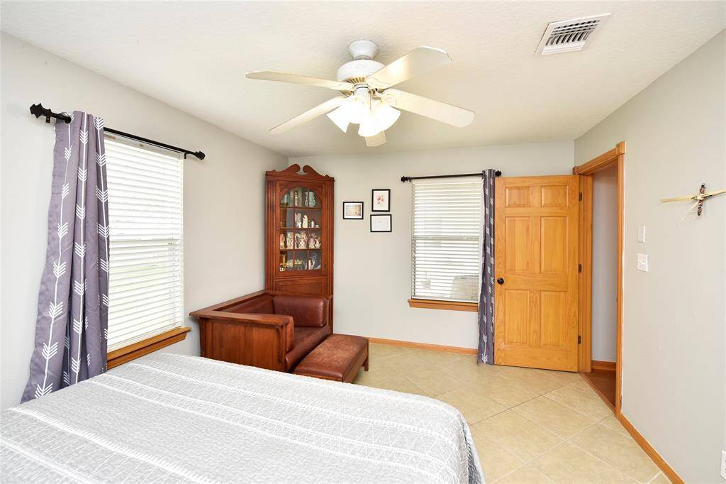 This bedroom features neutral tones with tiled flooring and ample natural light through two windows with patterned curtains. A ceiling fan provides comfort, and a wooden door and trim add warmth. A cozy seating area and display cabinet enhance the space.