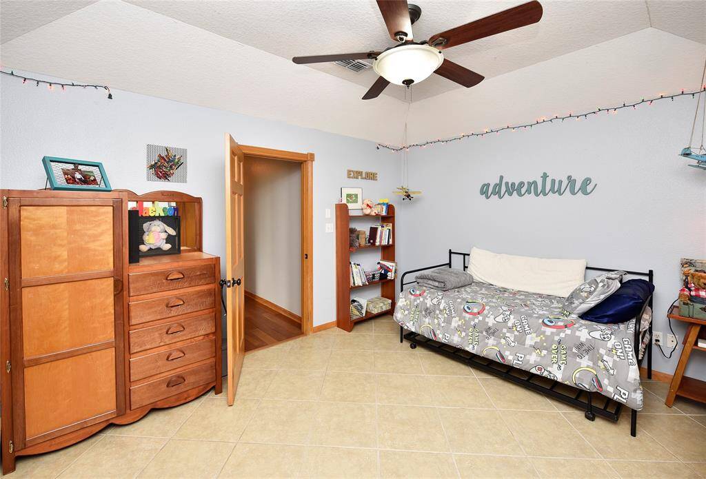 This room is a cozy, kid-friendly space with light blue walls and tiled flooring. It features a ceiling fan, a wooden dresser, a bookshelf, and a daybed with playful bedding. Decor includes string lights and an "adventure" wall sign, creating a warm and inviting atmosphere.