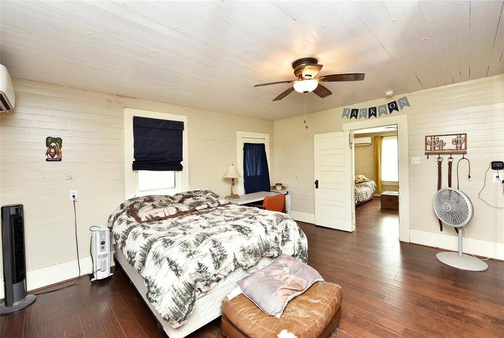 Spacious bedroom with wood floors and light-colored walls, featuring a ceiling fan and ample natural light. There's a cozy bed, a small desk, and an adjoining room visible through an open doorway.