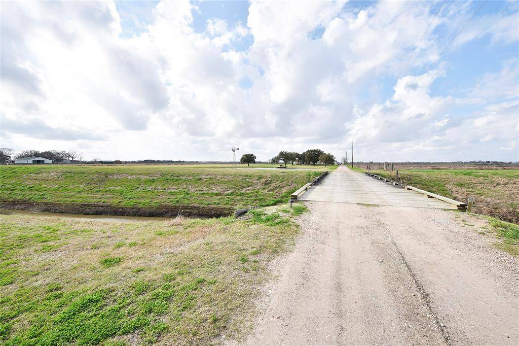 Expansive rural landscape featuring a long gravel driveway leading to a distant home, surrounded by open fields and under a partly cloudy sky, offering a peaceful, secluded setting.