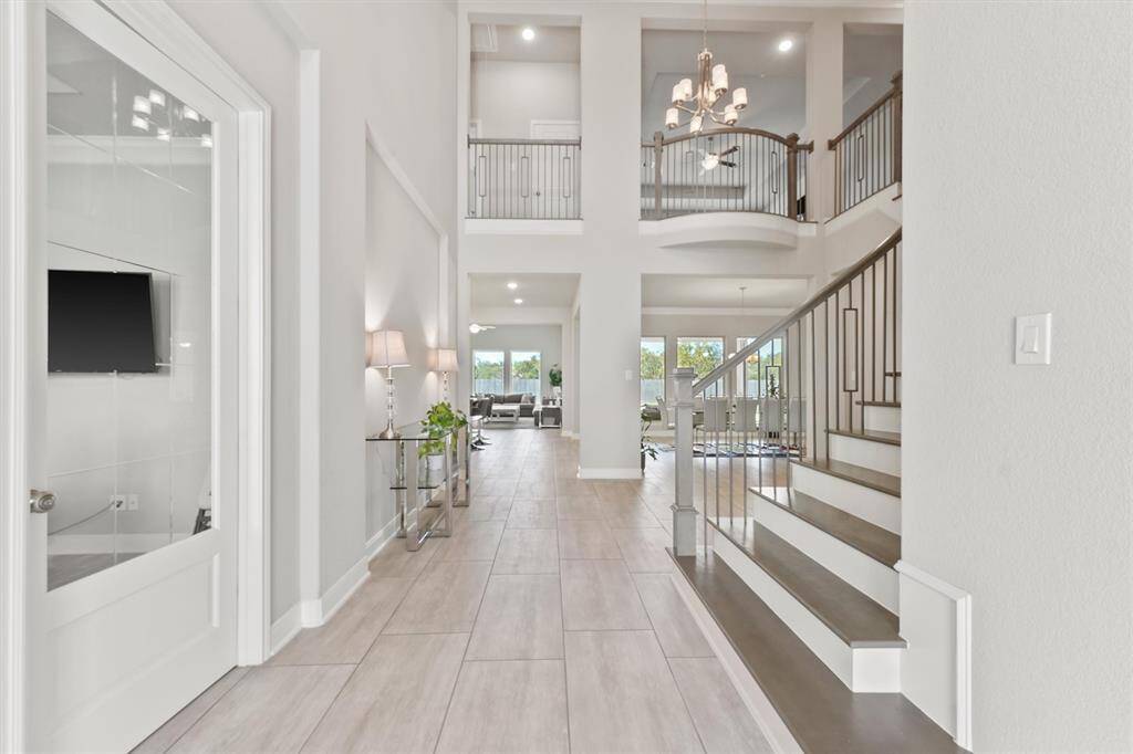 Modern, spacious entryway with high ceilings and an open layout featuring a sleek staircase with metal railings, elegant tile flooring, and abundant natural light. The area leads into a bright living space, with a view of the outdoors through large windows.