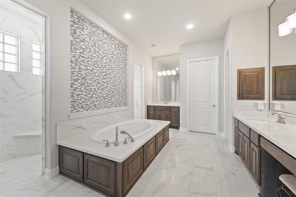This elegant bathroom features a spacious bathtub with a modern tile backsplash, dual sinks with ample counter space, and a separate shower. The sleek design includes marble-like flooring and warm wooden cabinetry, creating a luxurious and functional space.