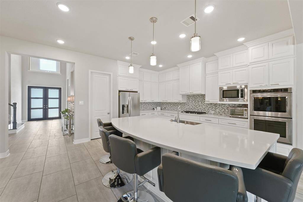 This modern kitchen features sleek white cabinetry, a large central island with seating, and stainless steel appliances. The space is illuminated by recessed lighting and pendant lights, while the open layout flows into a bright entryway with double doors.