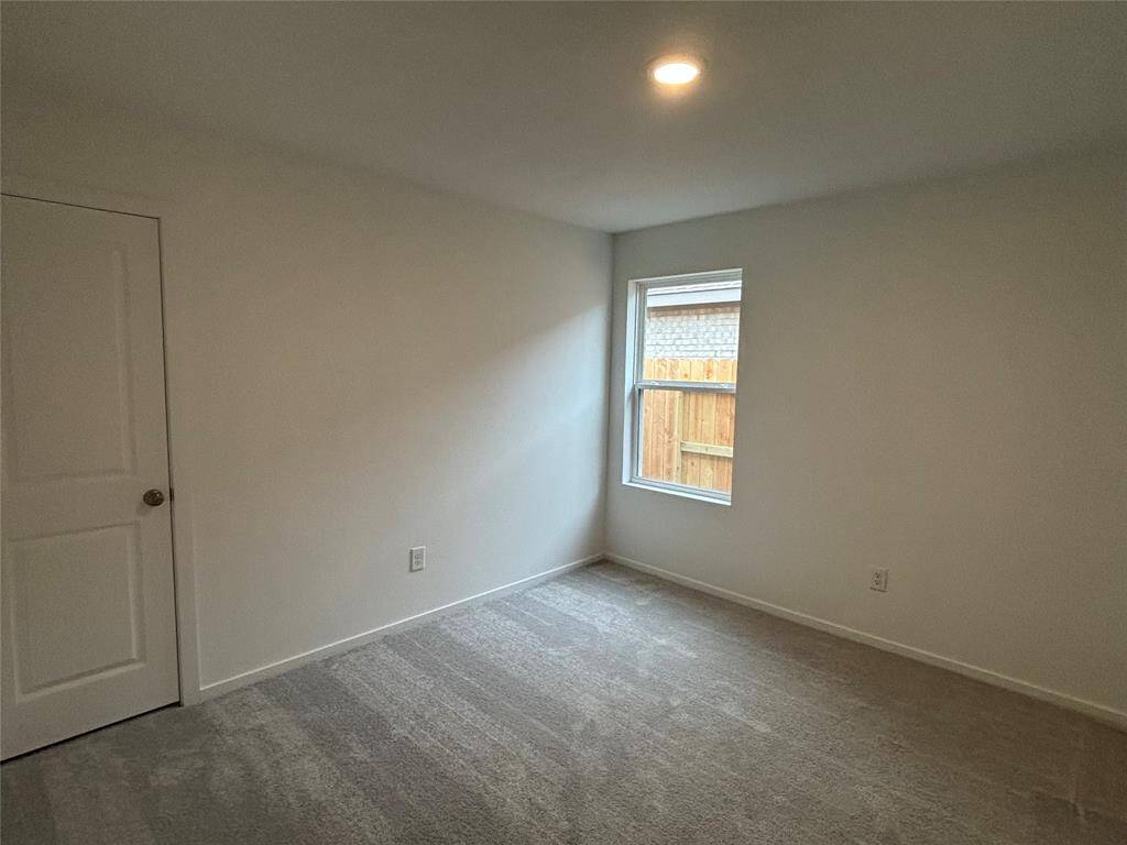 This photo shows a small, carpeted room with neutral walls, featuring a single window and a ceiling light. There's a closed door on one side, suggesting a cozy, minimalist space ideal for a bedroom or home office.