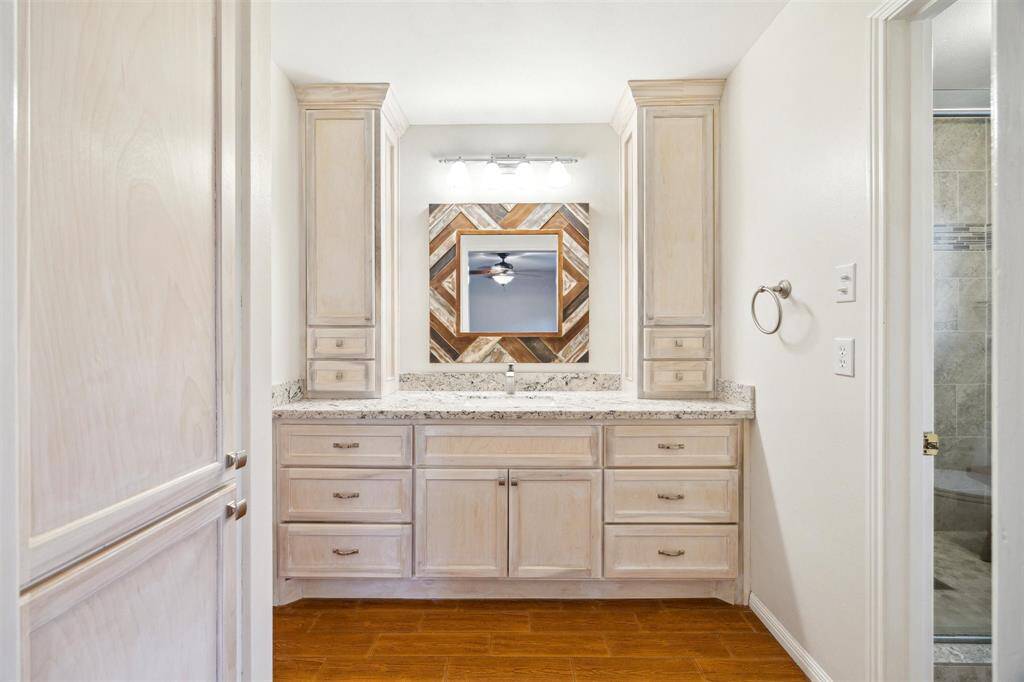 Bright, modern primary bathroom with ample storage. Patterned mirror above a granite countertop and light wood cabinetry. Adjacent to a tiled shower area.
