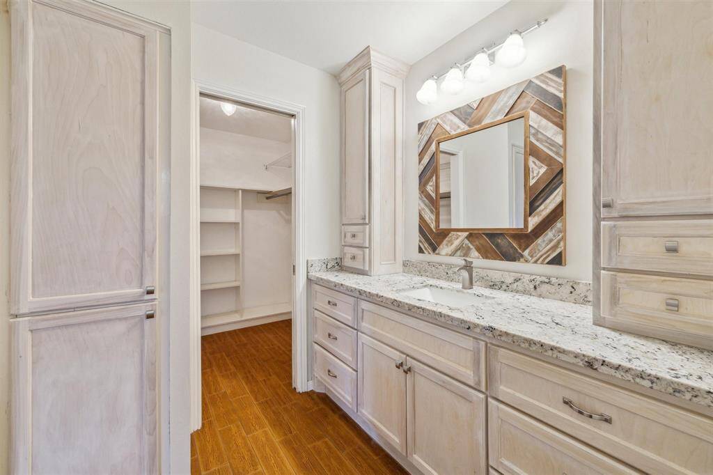 Primary bathroom with light wood cabinetry and a granite countertop. The space offers a large mirror with a decorative frame and modern lighting. An adjacent walk-in closet with shelving offers plenty of storage.