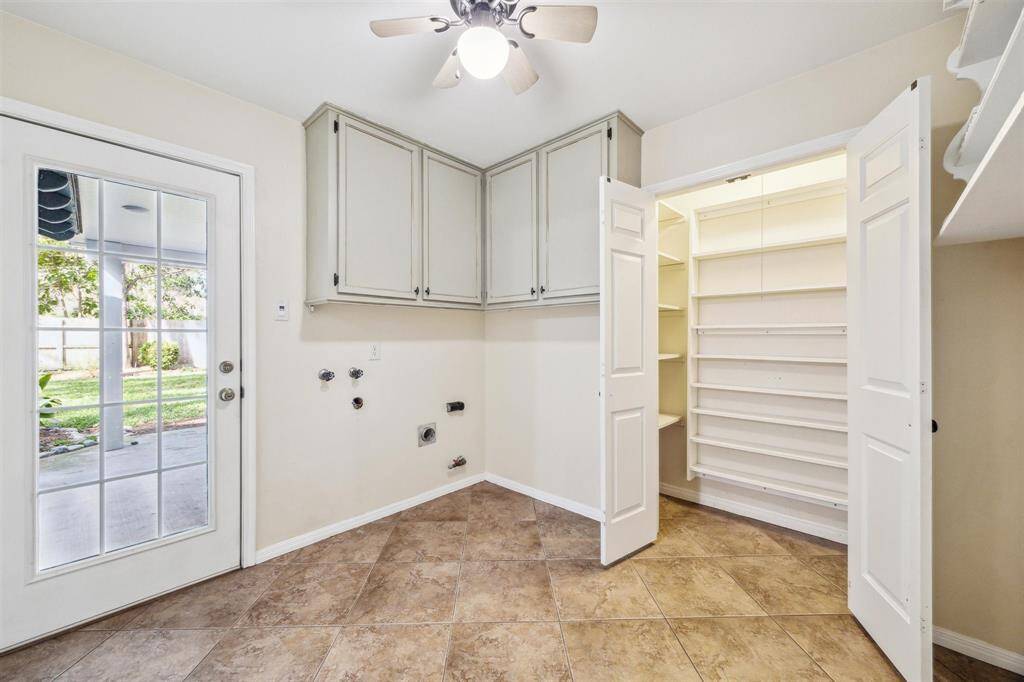 This utility room has tiled flooring, a ceiling fan, and ample natural light from a glass-paneled door leading to the backyard. It includes upper cabinets for storage, a spacious pantry, and hookups for laundry appliances.