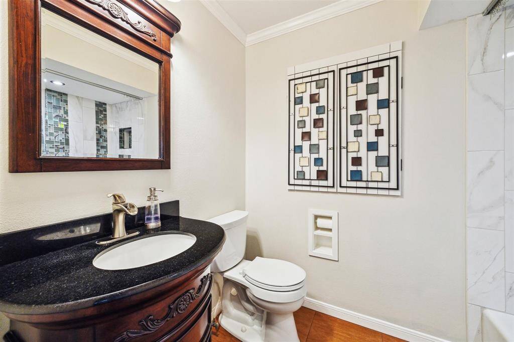 Hall bathroom offers decorative vanity with a dark countertop and an elegant wood-framed mirror. It has a modern toilet, decorative glass wall accents that offer more storage behind, and a tiled tub/shower combo.