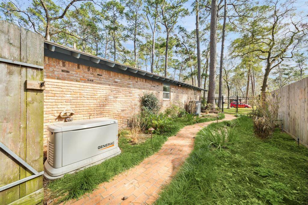 Side yard with a brick pathway, bordered by a wooden fence and a brick exterior wall. The area is fenced, offering privacy, and is surrounded by tall trees.