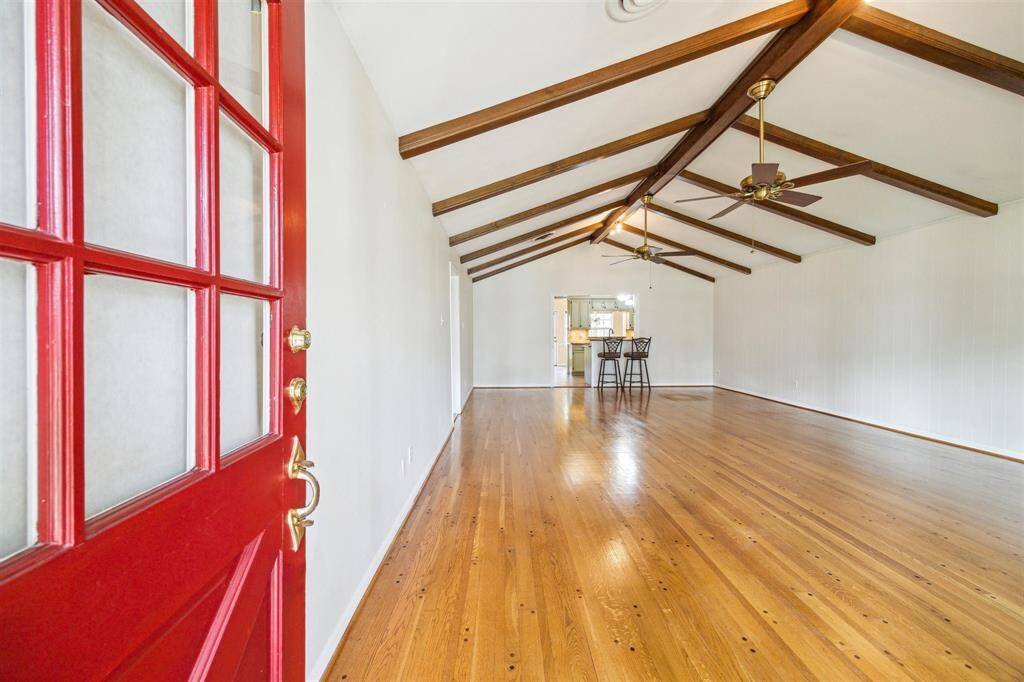 This photo shows off the oversized open-concept living area with beautiful hardwood floors and a vaulted ceiling with exposed wooden beams. The room is well-lit and includes ceiling fans for comfort. A cranberry front door adds a pop of color and character.