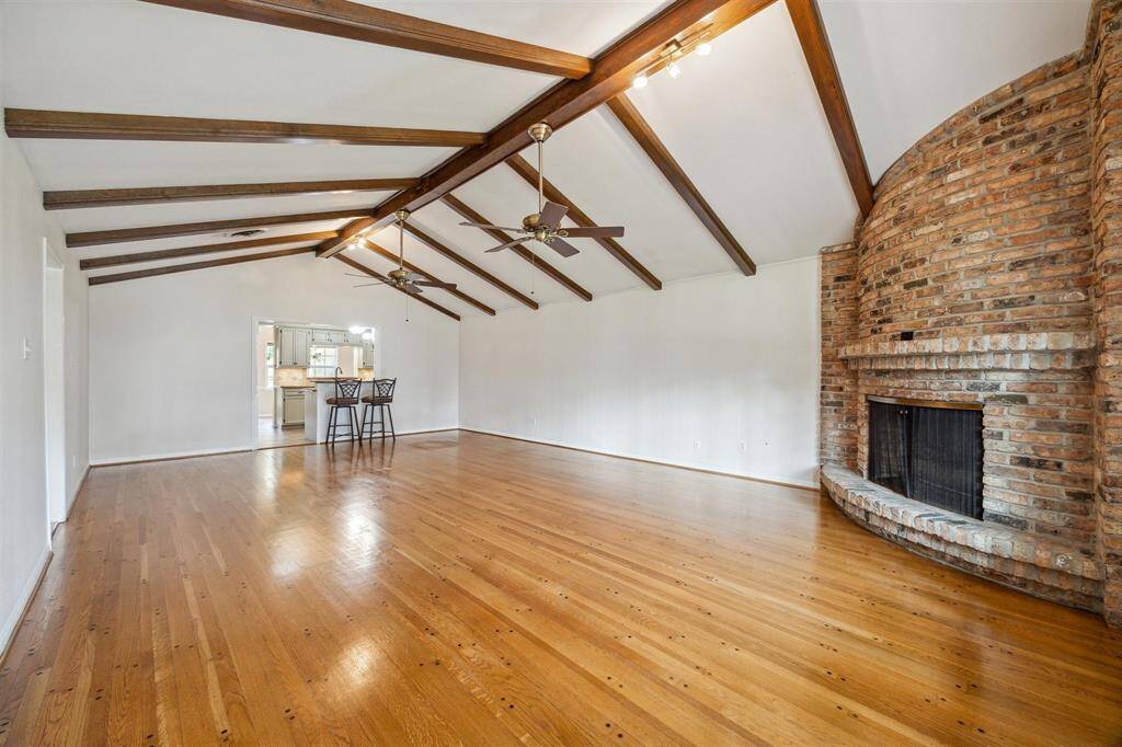 This family room has a vaulted ceiling with exposed wooden beams and a unique curved brick gas log fireplace. The hardwood floors add warmth, and there's an open view into the kitchen area, providing a great flow for entertaining.