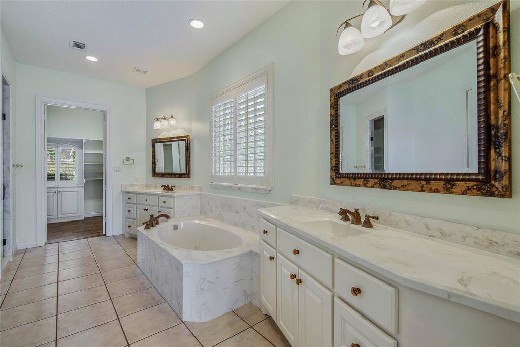 This primary ensuite bathroom features a spacious layout with a luxurious soaking tub, dual sinks with marble countertops, elegant mirrors, and ample storage. The room is brightened by natural light from a window with plantation shutters, highlighting the stylish tile flooring.