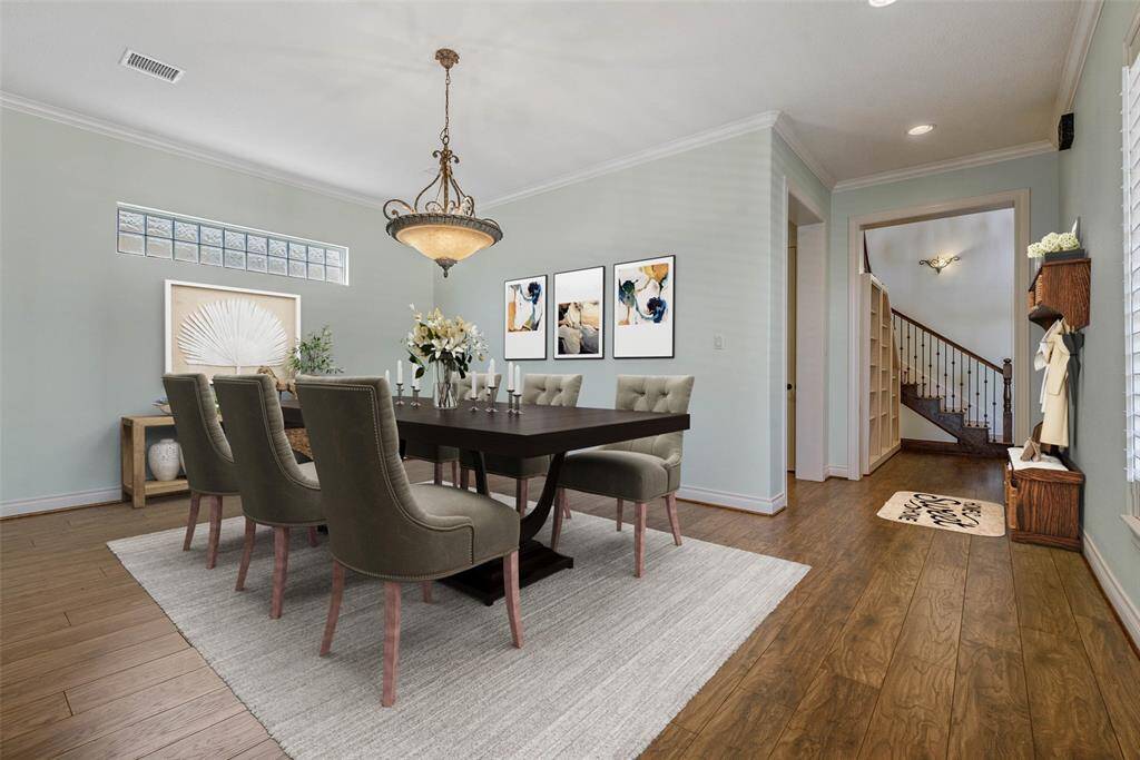 This virtual staged photo showcases an elegant dining room with a large table and upholstered chairs. It features hardwood floors, a stylish chandelier, and tasteful wall art. The room is connected to a hallway with a staircase, offering a welcoming, open layout.