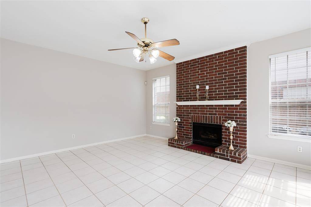 This bright living area features a cozy brick fireplace, large windows with blinds for natural light, a ceiling fan, and easy-to-maintain tiled flooring. Perfect for relaxation and gatherings.